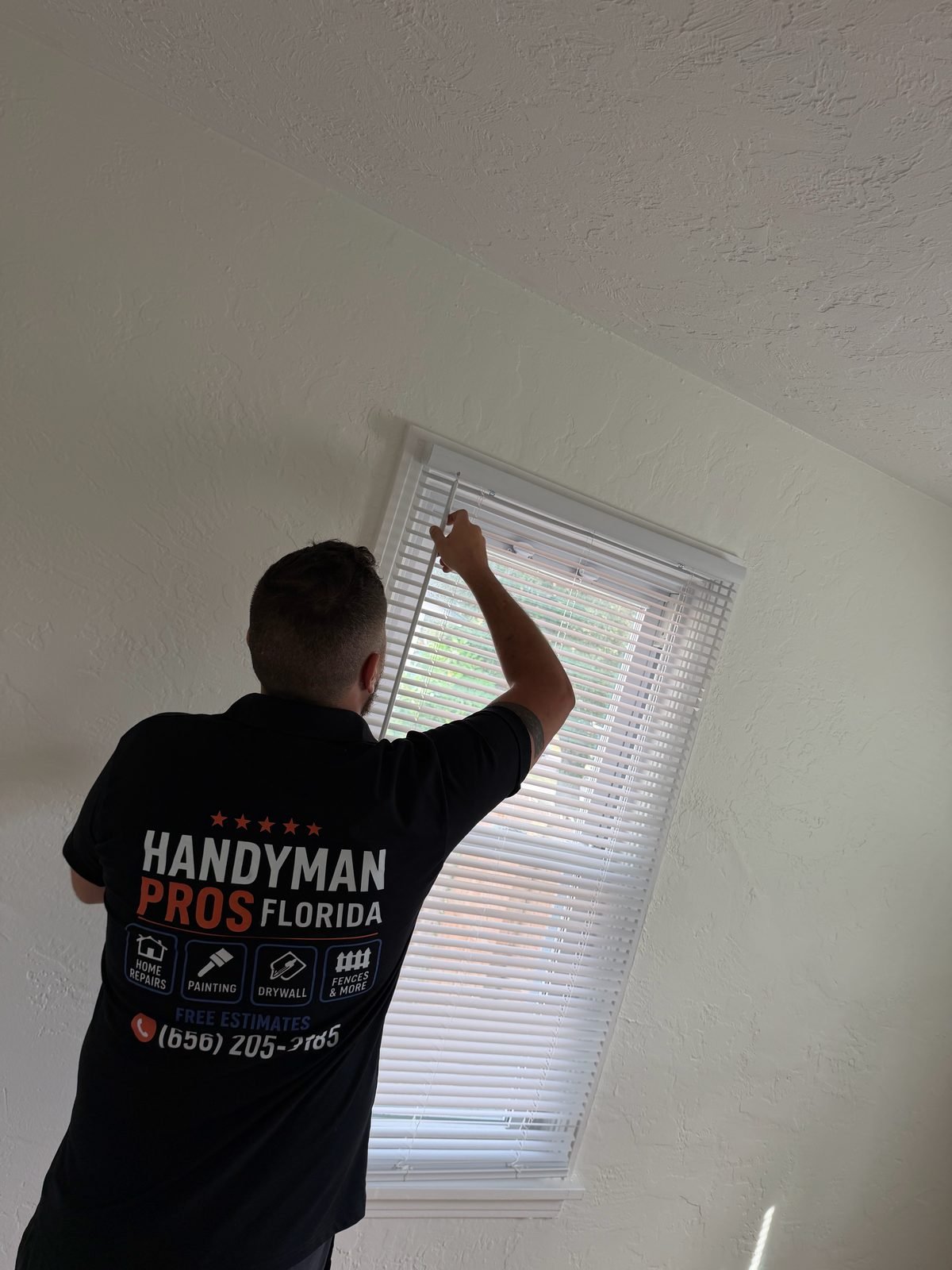 Handyman Pros Florida technician installing window blinds in a Tampa Bay home, branded shirt with phone (656) 205-3185